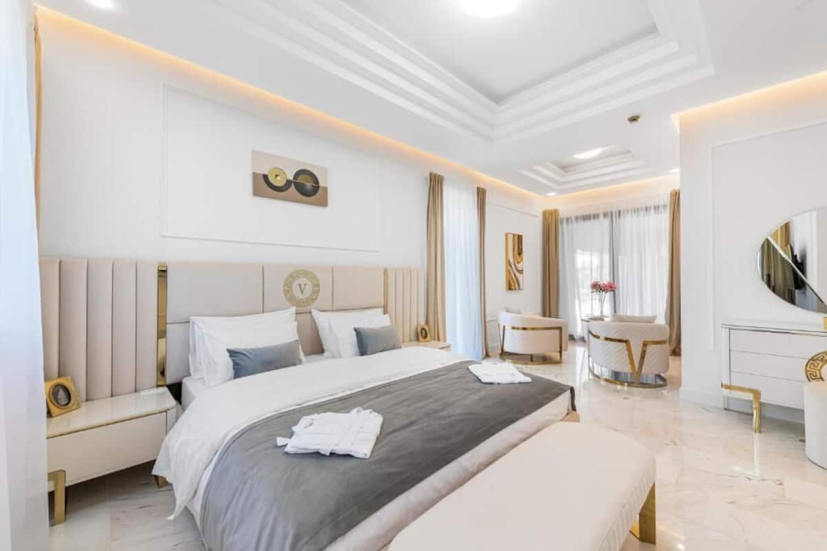 Una habitación de hotel moderna con una cama grande, mesitas de noche, un tocador con espejo y una zona de estar con una silla y una mesita.