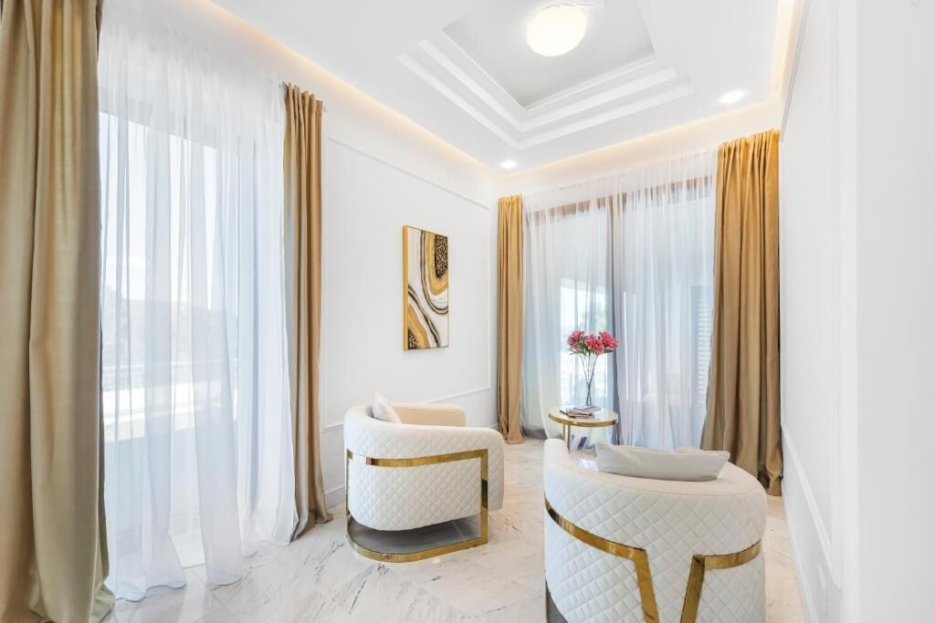 A modern hotel room with a marble floor, two white armchairs with gold accents, a round ottoman, a small table with a vase of flowers, and sheer curtains.