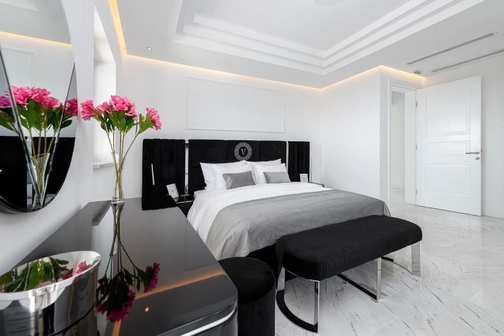 A modern bedroom with a large bed, a bedside table with a vase of flowers, a mirror, and a bench.