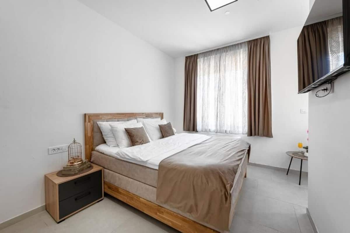 Un dormitorio con cama, mesita de noche, televisor montado en la pared y ventana con cortinas.