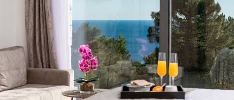 A bedroom with a large window, a bed, a bedside table with a tray of orange juice and glasses, and a potted orchid.