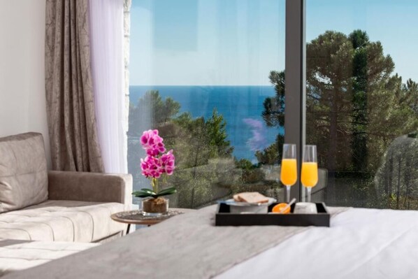 Une chambre avec une grande fenêtre, un lit, une table de chevet sur laquelle se trouve un plateau avec du jus d’orange et des verres, et une orchidée en pot.