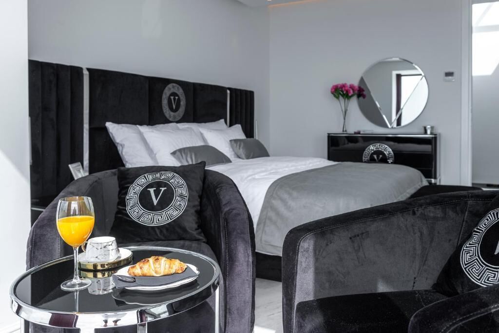 A modern hotel room with a bed, a round mirror, a bedside table with breakfast, and matching black upholstered chairs.