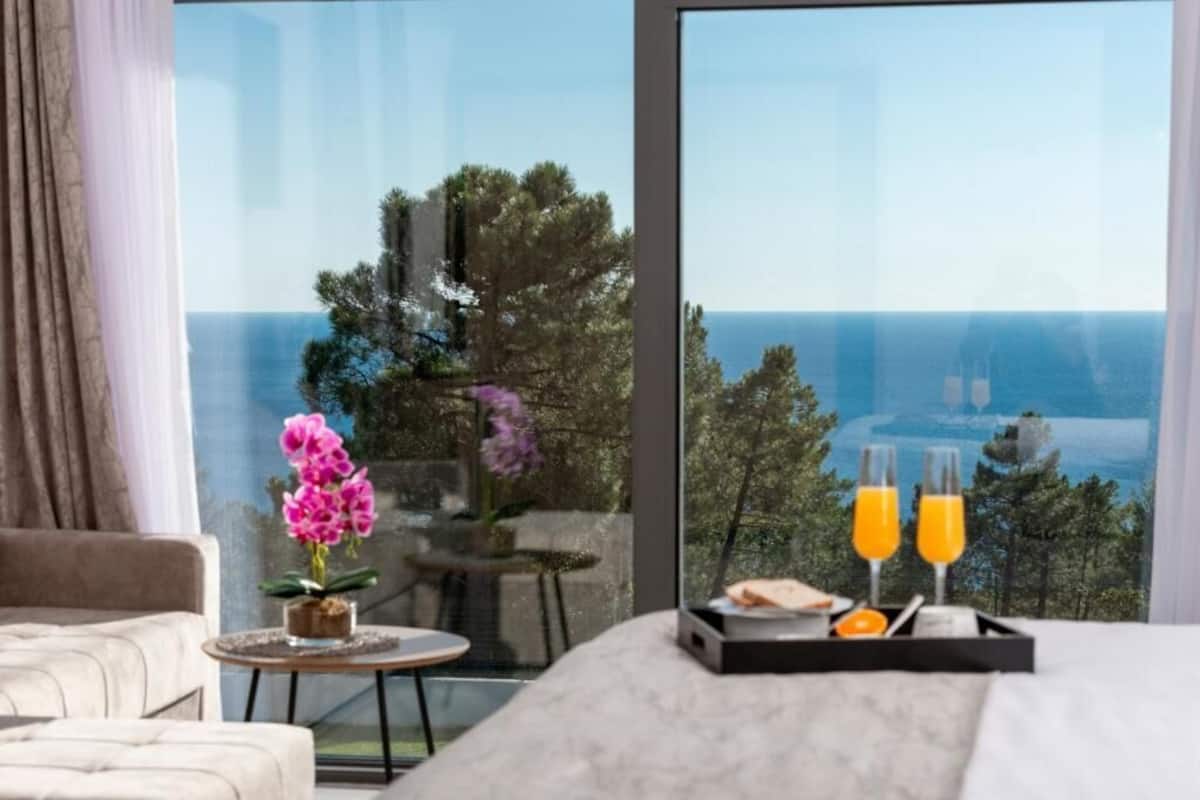 Una habitación de hotel con un ventanal grande, una cama, una pequeña mesa redonda con una orquídea en maceta y una bandeja con dos vasos de jugo de naranja.