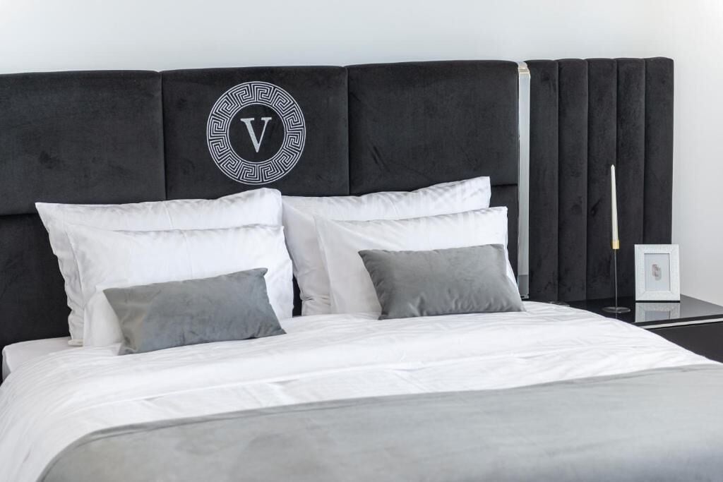 Una cama con ropa de cama blanca y gris, un cabecero negro con un logotipo circular y una mesita de noche con una lámpara y un marco de fotos.