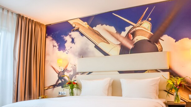 Et hotelværelse med en stor seng, et natbord med en lampe, en vase med blomster og et muralmaleri af en helikopter på væggen.