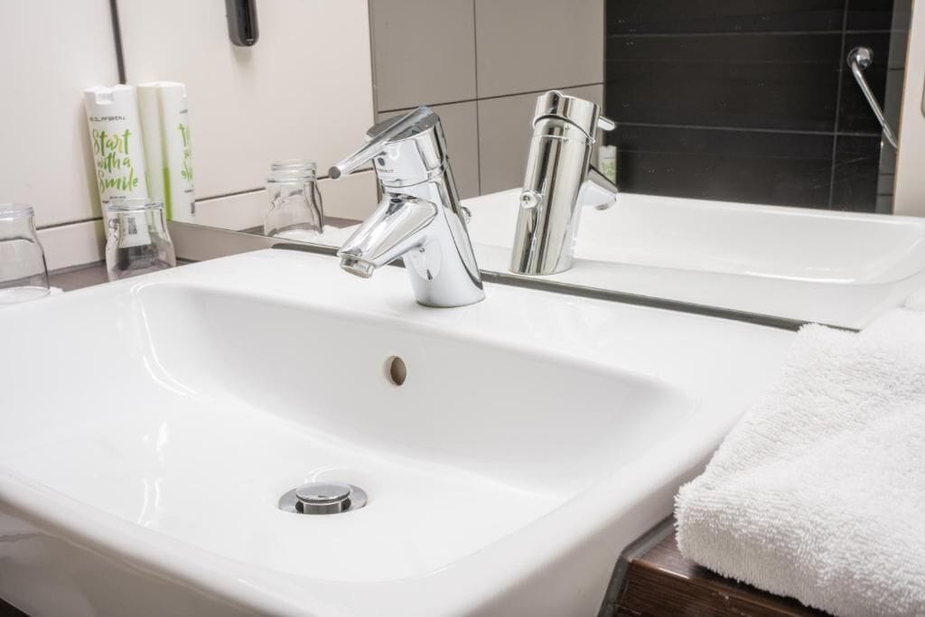 Un lavabo blanc dans une salle de bain, avec des robinets chromés, un miroir et une serviette blanche posée sur une surface en bois.