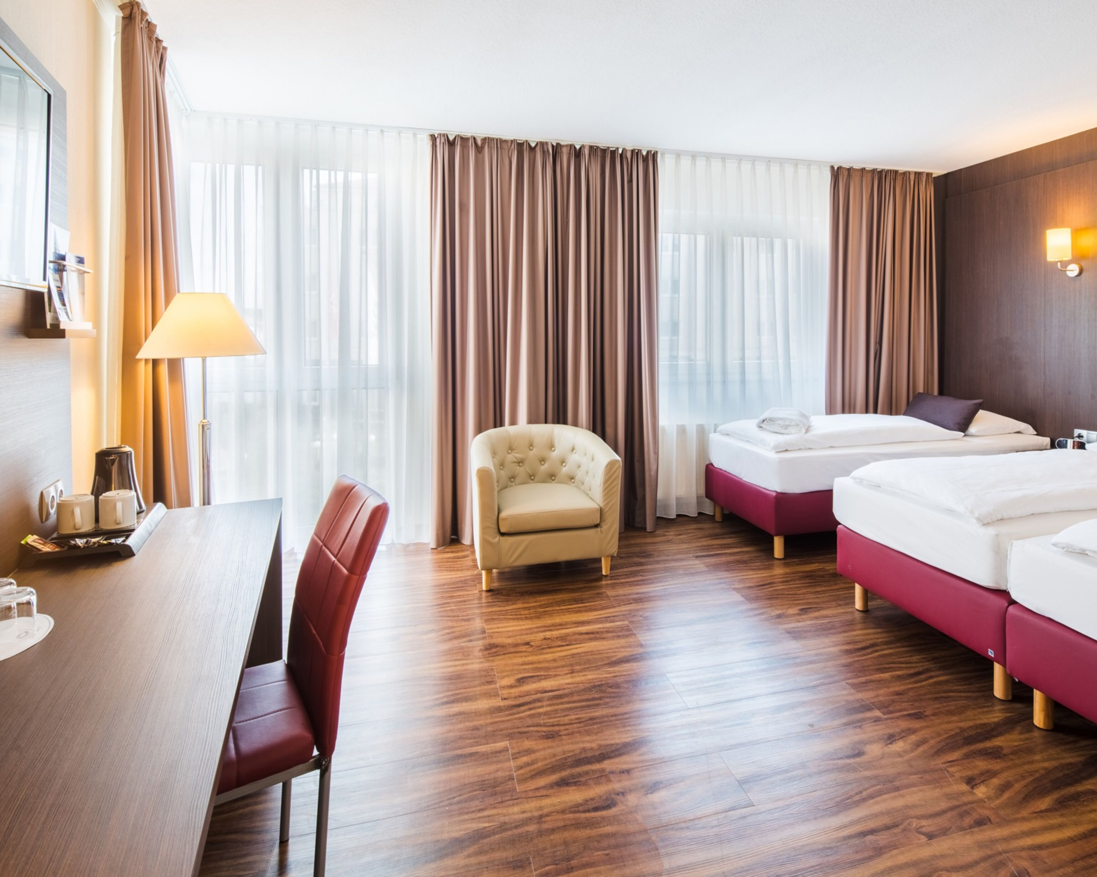 Habitación de hotel con suelo de madera, un escritorio con lámpara, una silla, dos camas y un ventanal con cortinas.