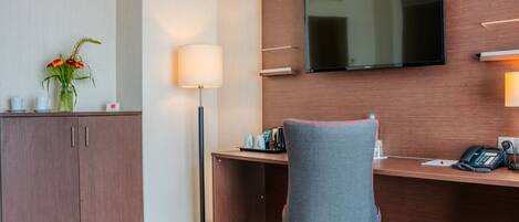 A hotel room with a wooden desk, a flat-screen TV mounted on the wall, a chair, a lamp, a cabinet, and a vase with flowers.