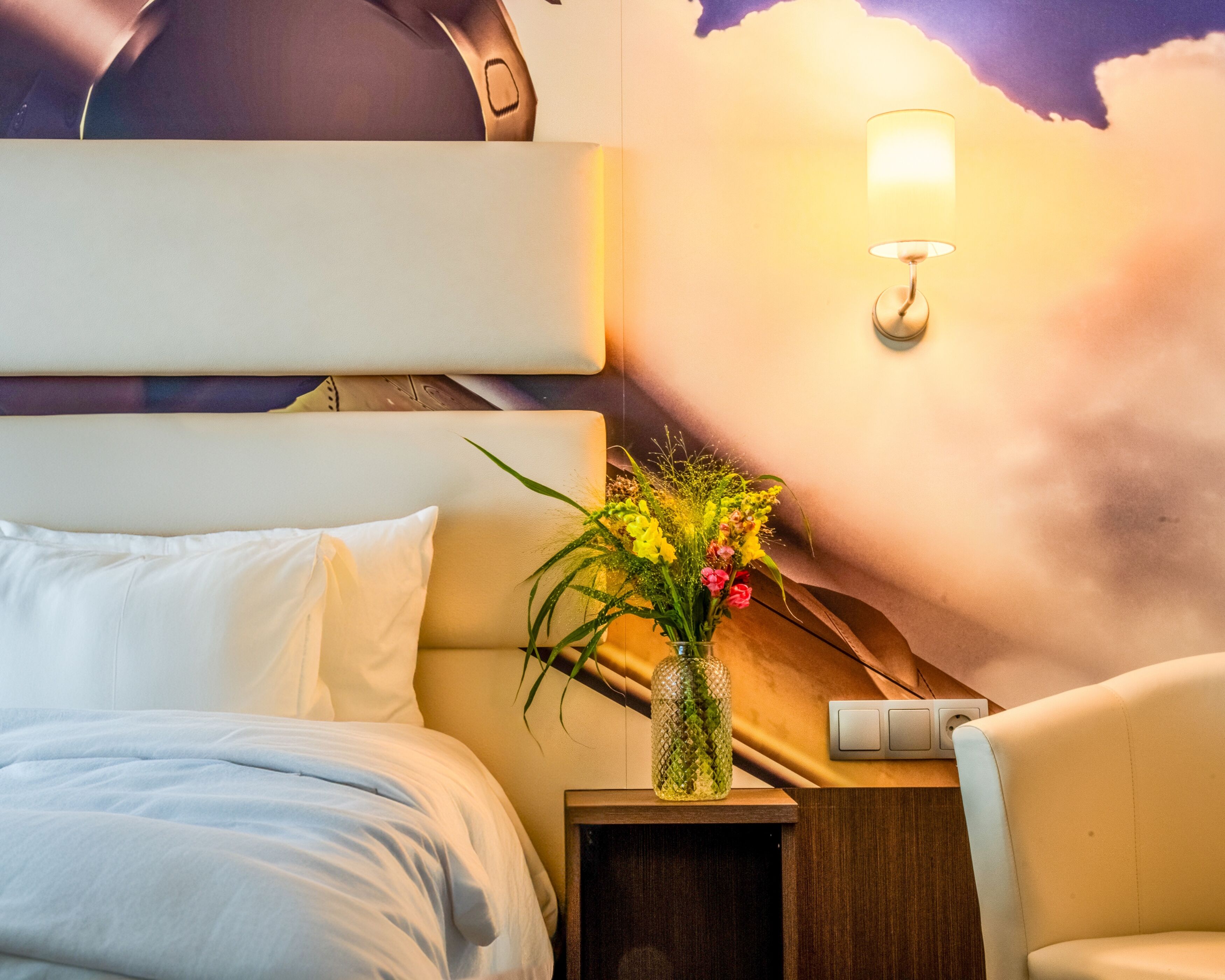 A hotel room with a bed, a nightstand with a vase of flowers, a lamp, and a wall-mounted light.