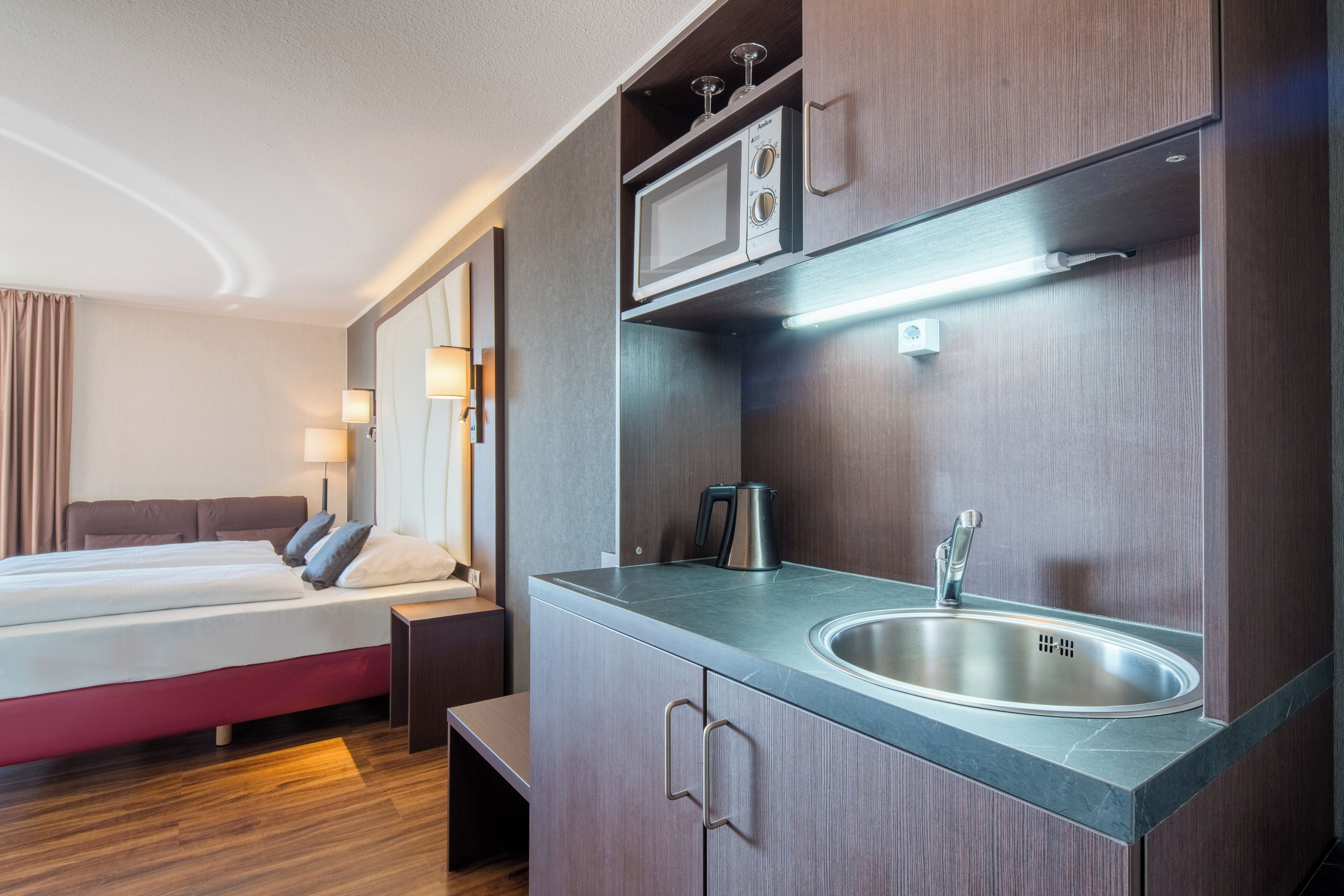 Ein modernes Hotelzimmer mit Bett, Nachttisch, Lampe und einer integrierten Küchenzeile mit Mikrowelle, Wasserkocher und Spüle.