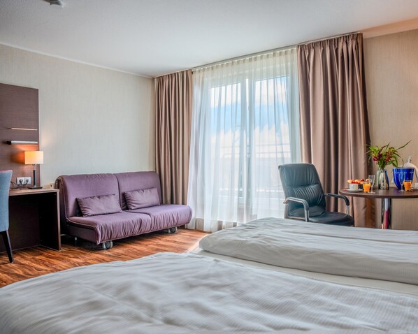 Habitación de hotel con cama, sofá, escritorio con lámpara, una silla y ventana con cortinas.