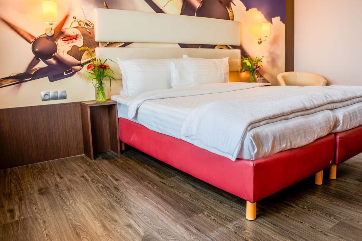 Quarto moderno com estrutura de cama vermelha, roupa de cama branca, mesa de cabeceira com abajur, relógio fixado na parede e um objeto decorativo na parede.