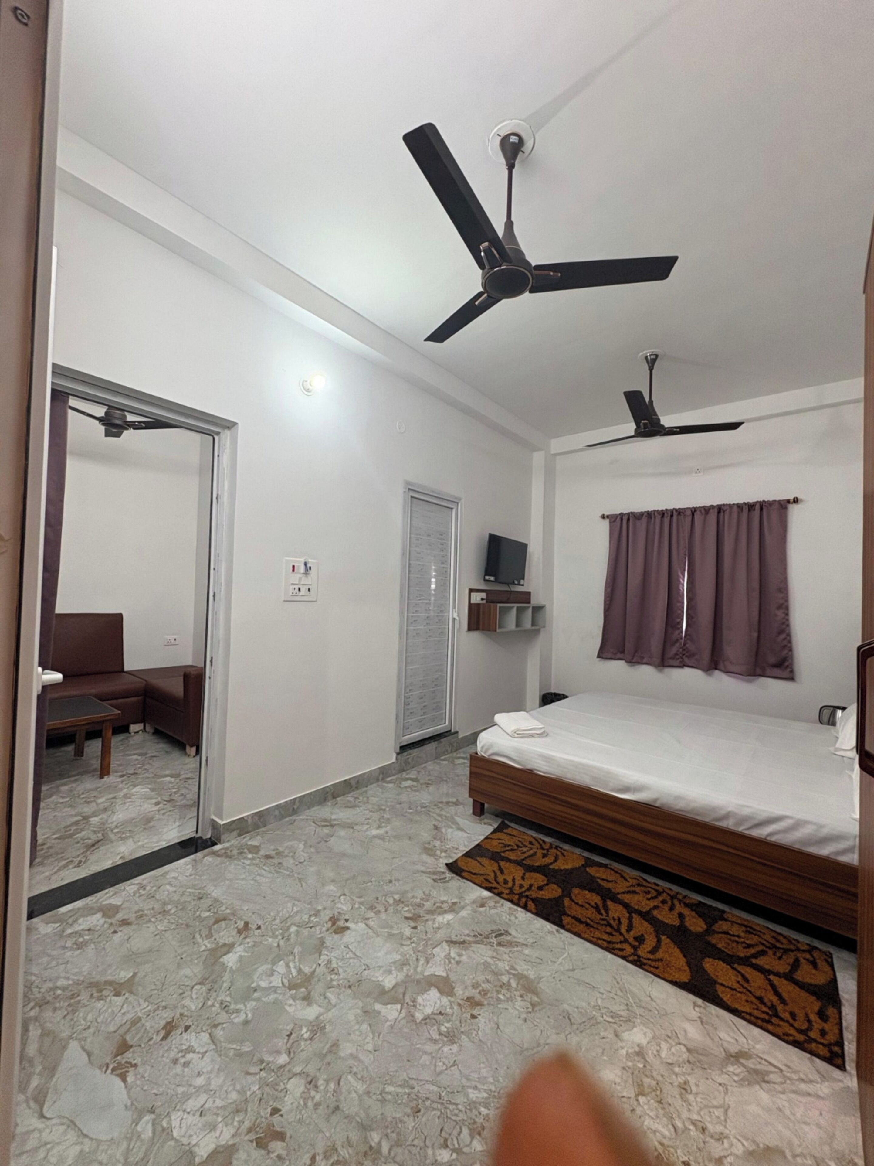A bedroom with a bed, two ceiling fans, a television on a shelf, a door, and a patterned rug.