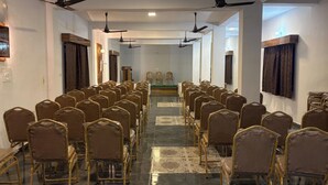 Un salón de conferencias con filas de sillas, ventiladores de techo y un atril al frente.