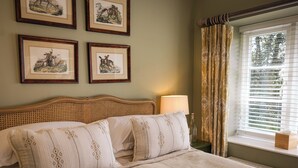 Ein Bett mit Kopfteil, eine Nachttischlampe, ein Fenster mit Jalousien und gerahmte Bilder an der Wand.