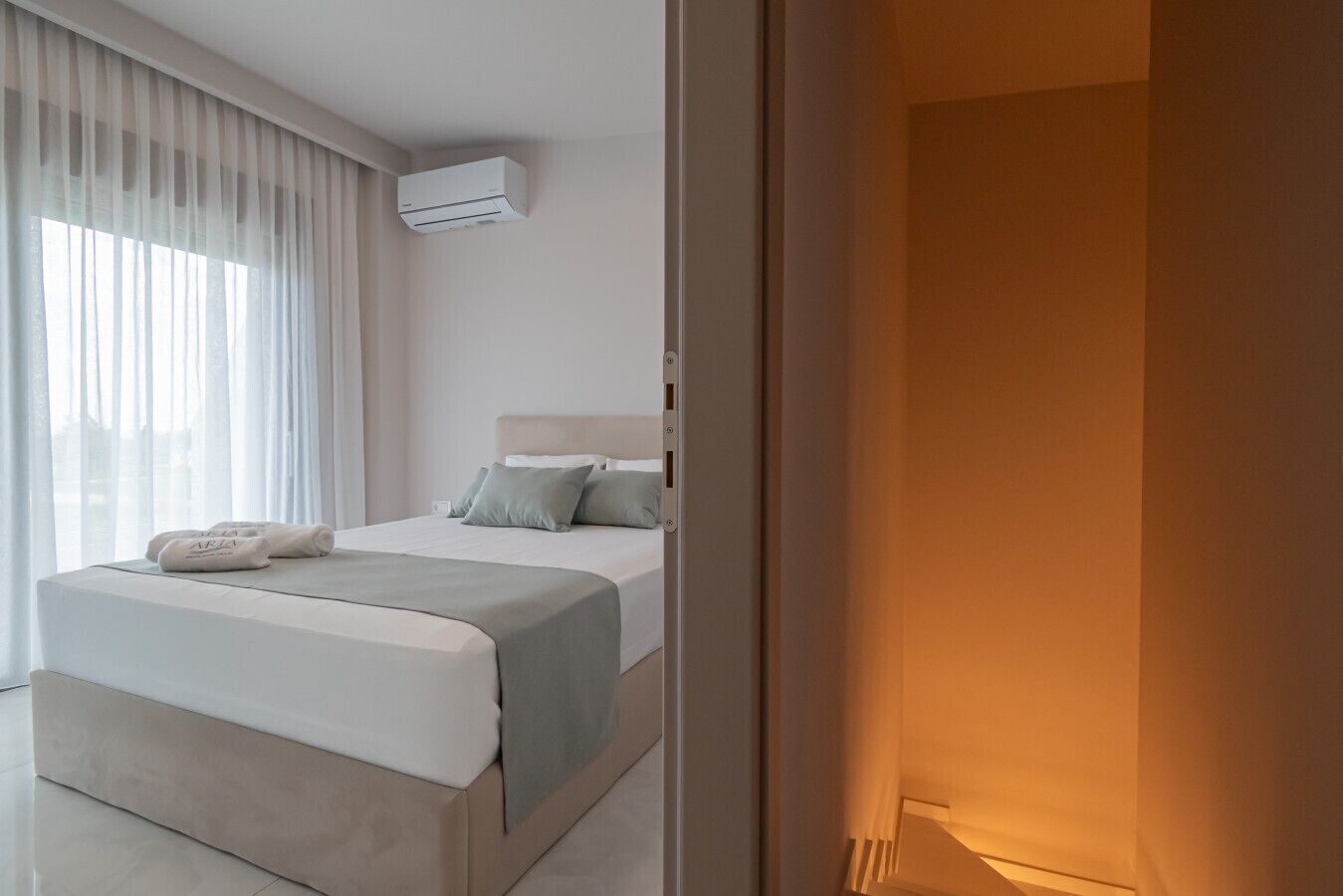 A hotel room with a bed, a bedside table, a window with curtains, and an air conditioning unit.