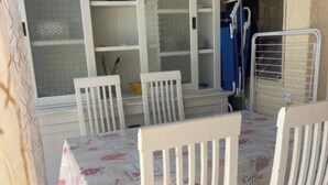 A dining area with a table covered by a tablecloth, white chairs, a cabinet with glass doors, and a metal rack. - 88 Heliovillage (Agde)