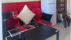 A living room with a red and black sofa, a small table, a white refrigerator, a wall-mounted air conditioner, a staircase, and a hanging light fixture. - 88 Heliovillage (Agde)