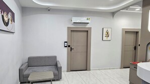 A modern hotel room with a grey sofa, a small table, a door, an air conditioning unit, and a framed artwork on the wall. - Hotel HR Comfort (Ahmedabad)