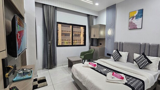 A modern hotel room with a bed, bedside table, chair, TV, artwork, and a window with curtains. - Hotel HR Comfort (Ahmedabad)
