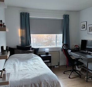 Ein Schlafzimmer mit einem Bett, einem Schreibtisch mit Computer, einem Stuhl, einer Heizung und einem Fenster mit Vorhängen.