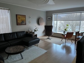 Ein modernes Wohnzimmer mit einem schwarzen Ledersofa, einem runden Couchtisch, einer hohen Stehlampe und Blick auf Bäume durch ein großes Fenster.