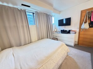 Ein Schlafzimmer mit einem Bett, einem an der Wand montierten Fernseher, einem Schreibtisch mit Stuhl und einer Tür, an der eine Tasche hängt.