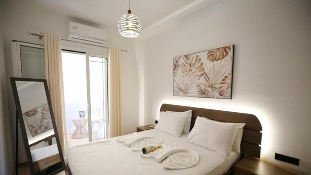 A hotel room with a bed, a bedside table, a nightstand, a mirror, a window with curtains, and a wall-mounted artwork. - Premium Luxury Living Lefkada (Lefkada)