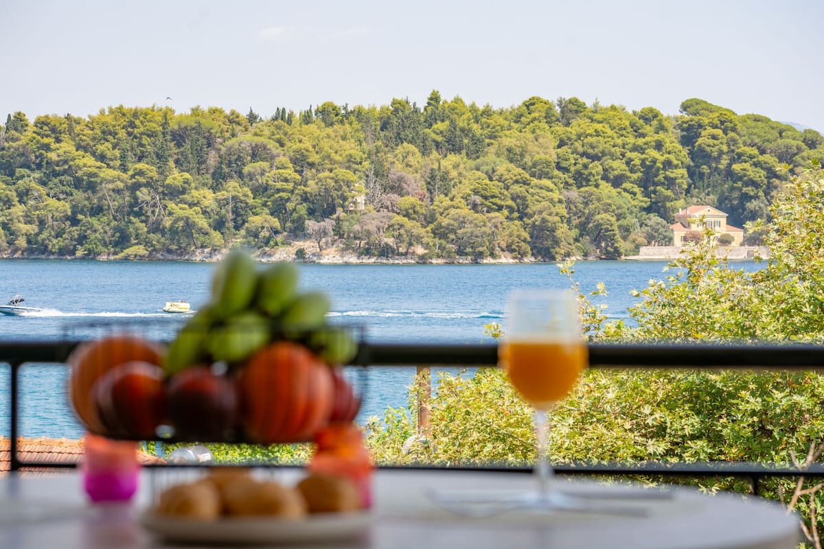 A table with fruit and a glass of wine, overlooking a coastal landscape with trees and a building.
