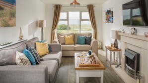 Ein Wohnzimmer mit einer Couch, einem Couchtisch mit Schachfiguren, einem Kamin, einem Fernseher und einem großen Fenster mit Vorhängen.