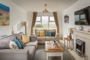 Ein Wohnzimmer mit einer Couch, einem Couchtisch mit Schachfiguren, einem Kamin, einem Fernseher und einem großen Fenster mit Vorhängen.