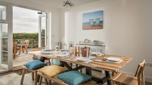 Ein Essbereich mit Holztisch, gedeckter für vier Personen, Bänken mit Kissen, Blick durch eine Glastür und Strandkunst an der Wand.