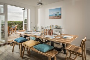 Ein Essbereich mit Holztisch, gedeckter für vier Personen, Bänken mit Kissen, Blick durch eine Glastür und Strandkunst an der Wand.