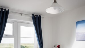 Ein Schlafzimmer mit Bett, Nachttisch, Lampe, einem Fenster mit Vorhängen und Blick ins Freie.
