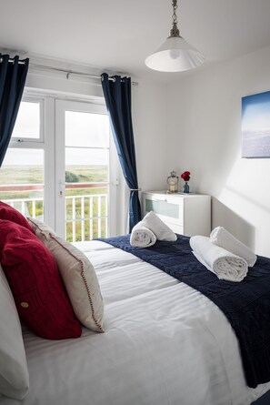 Ein Schlafzimmer mit Bett, Nachttisch, Lampe, einem Fenster mit Vorhängen und Blick ins Freie.