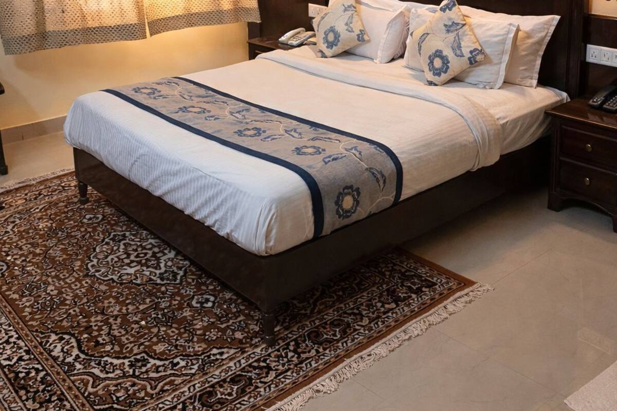 A neatly made bed with a patterned bedspread, a large painting of a tree by a lake on the wall, and a patterned rug on the floor.