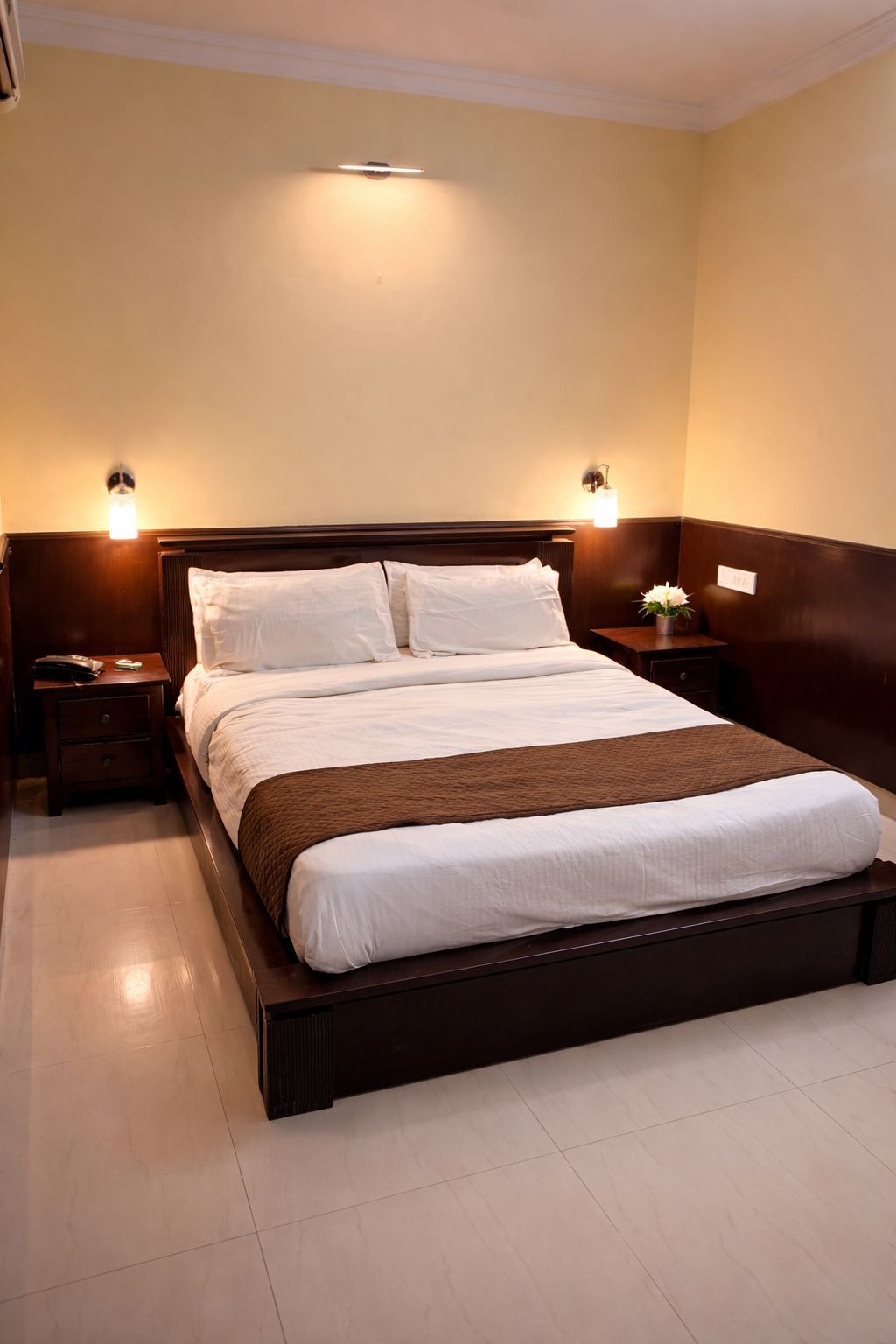 A hotel room with a large bed, two bedside tables, a nightstand, a wall-mounted light fixture, and a small table with a vase.
