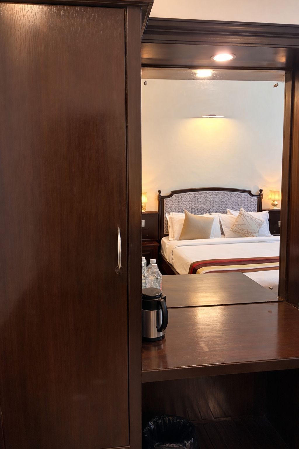 Habitación de hotel con una cama, lámparas de noche, un escritorio de madera, un cubo de basura y una botella de agua.
