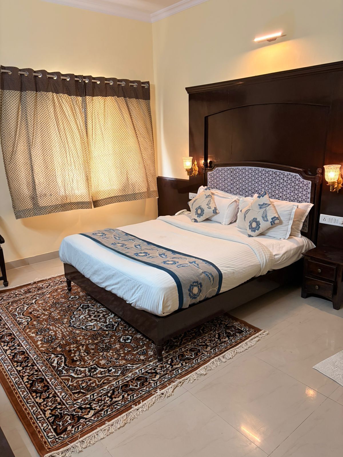 A bedroom with a bed, patterned curtains, a headboard, bedside lamps, and a patterned rug.