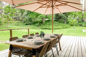 Eine Holzterrasse mit einem für sechs Personen gedeckten Tisch, umgeben von Korbstühlen, unter einem großen Sonnenschirm.