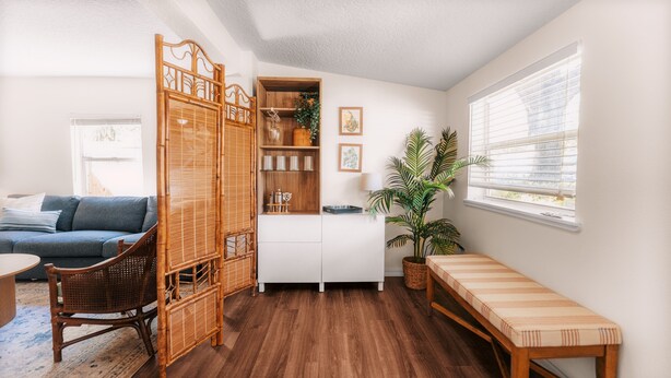A living room with a sofa, a wooden bench, a dining table, a bamboo partition, a potted plant, and a window with blinds.