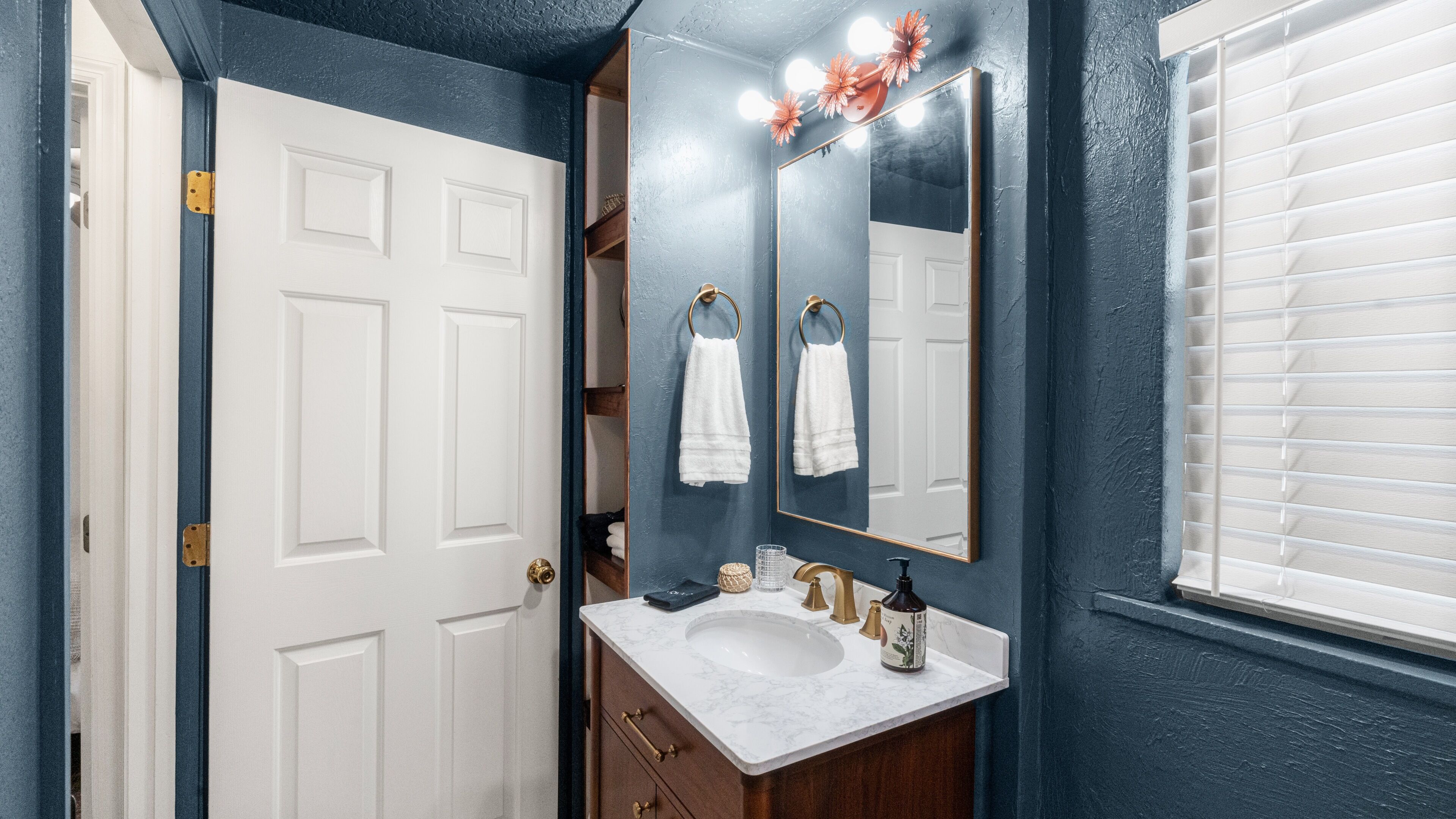 A bathroom with a white door, a sink with a marble countertop, a mirror with a gold frame, and a window with blinds.