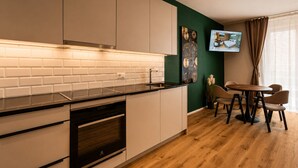 Una cocina moderna con armarios blancos, encimera negra y un horno. Hay un comedor con mesa y sillas, y un televisor montado en la pared.