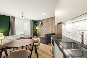 Eine moderne Küche mit schwarzem Arbeitsplatz und Spüle aus Edelstahl, ein Essbereich mit rundem Tisch und Stühlen sowie ein Wohnbereich mit Sofa und Wand-Fernseher.