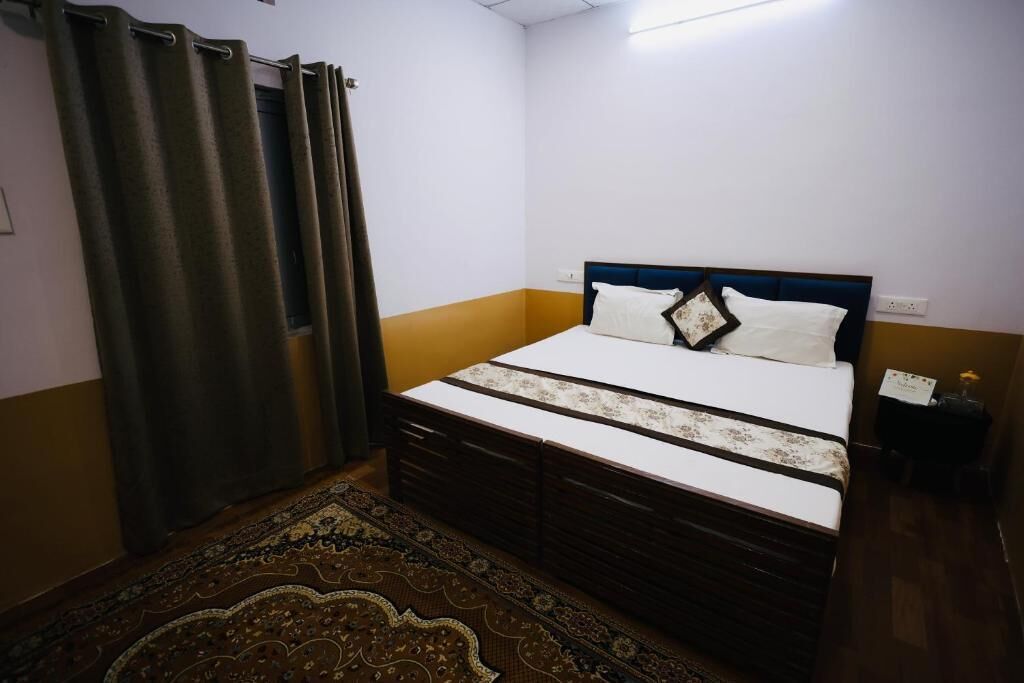 Un dormitorio con cama, mesita de noche, lámpara, ventana con cortinas y una alfombra estampada.