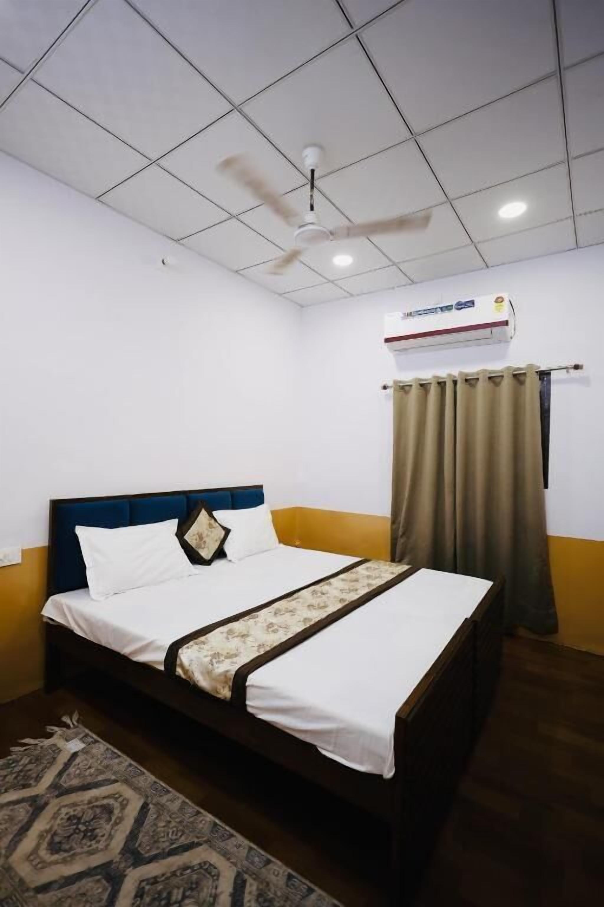 A hotel room with a bed, a ceiling fan, an air conditioning unit, and a window with curtains.
