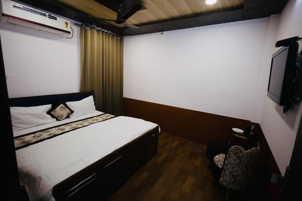 Um quarto com cama, ventilador de teto, ar condicionado, uma mesinha com cadeira e uma televisão na parede.