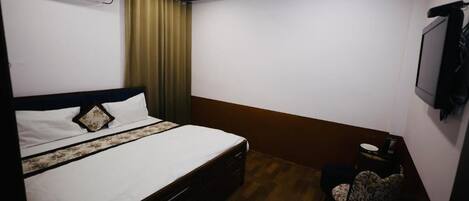 Um quarto com cama, ventilador de teto, ar condicionado, uma mesinha com cadeira e uma televisão na parede.
