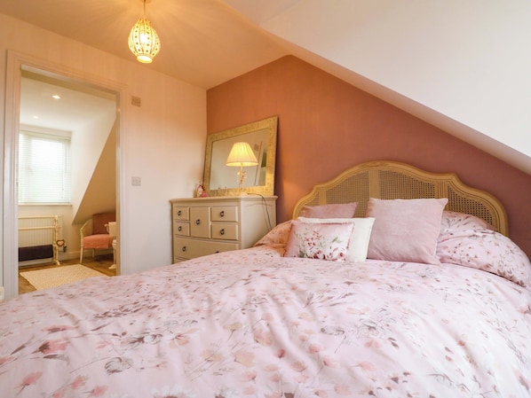 A bedroom with a sloped ceiling, a bed with floral bedding, a dresser, a mirror, and a hanging light fixture.